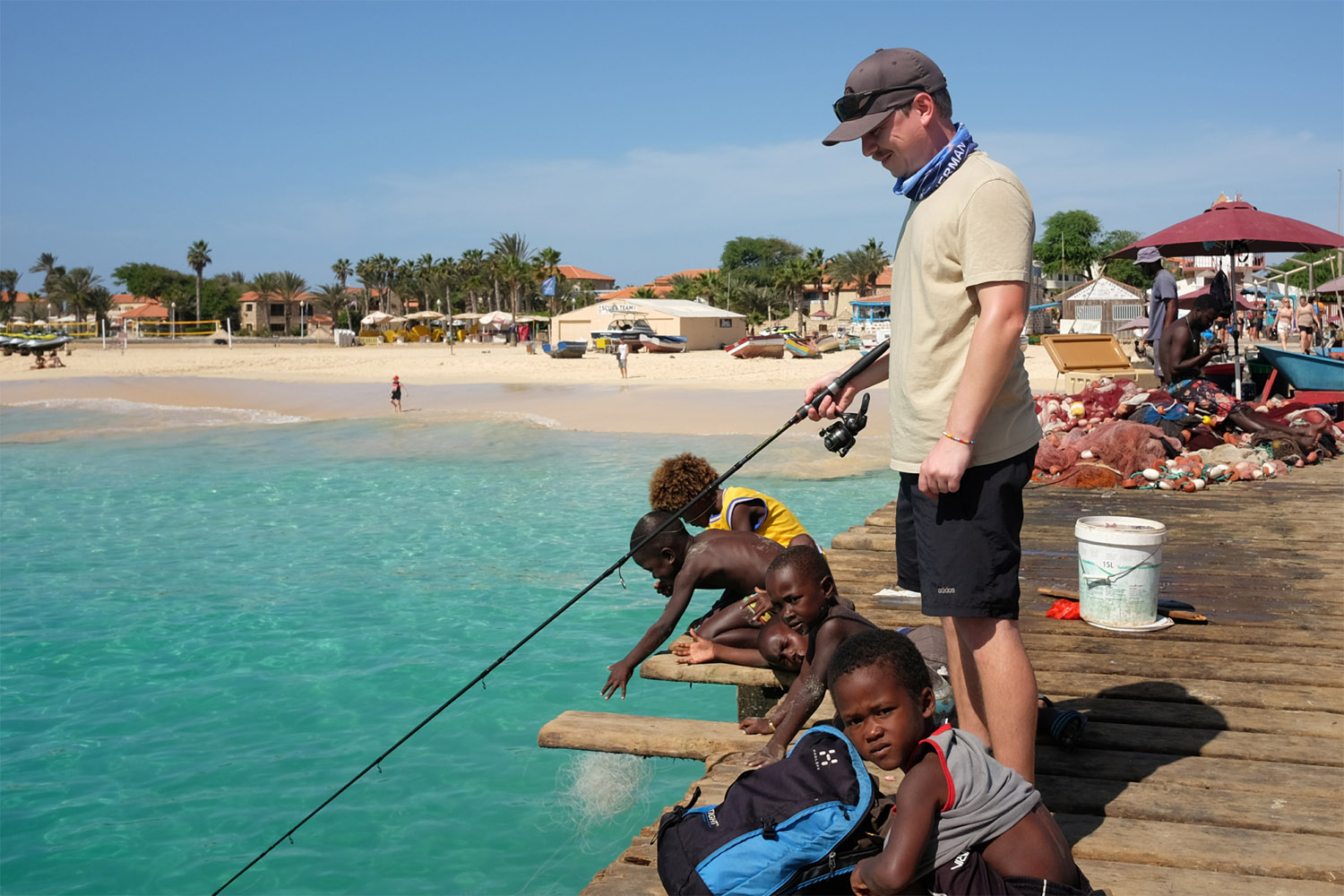 A man and children fishing from a pier in Cabo Verde, with a beach and resort in the background. 